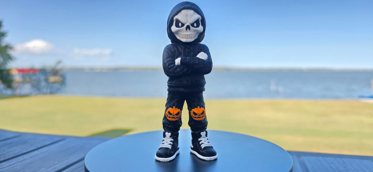 A small skeleton figurine wearing a black hoodie, black pants with orange pumpkin graphics, and black sneakers.